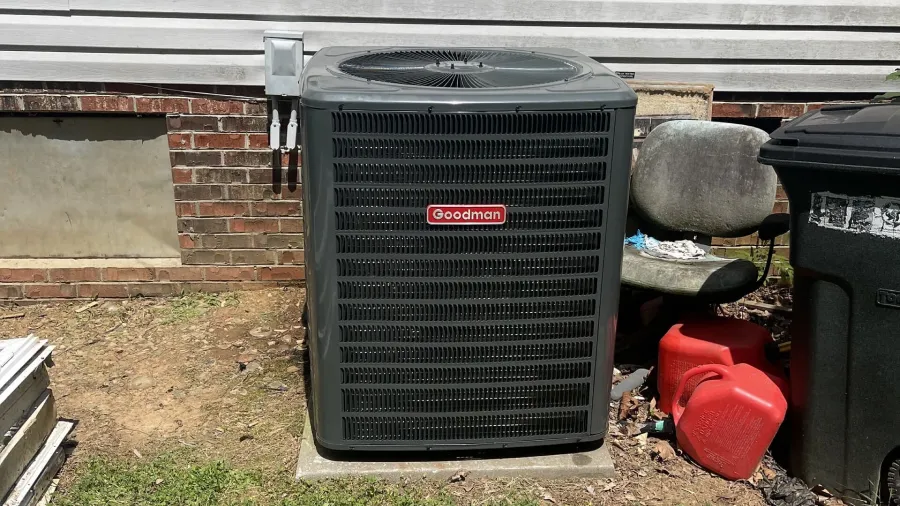 hvac repair services local A gray Goodman air conditioner unit with a red logo on a concrete slab sits next to a house wall and trash cans.